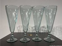 Four tall clear hand blown glasses shown side by side on a flat surface.