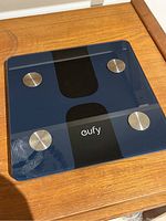 Top view of Eufy Smart Scale C1 showing black glass surface, four round metal sensors, and digital display area.