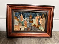 Framed ceramic artwork showing multi-colored buildings and towers in a stylized design with blue sky background
