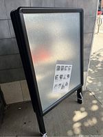 Photo of the folding sandwich board standing outdoors, showing its angled side profile and black frame with textured reflective display surface.