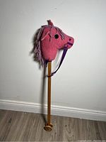Side view of a pink fabric hobby horse head on wooden stick with wheels on floor against white wall.