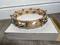 Side angle photo showing light wood circular tambourine frame with metal jingles, resting on white cardboard box.