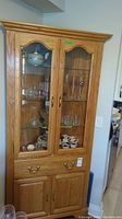 Front view of large wooden corner display cabinet showing upper glass inset doors with shelves containing glassware and teacups, small drawer in middle, and bottom wood cabinet doors.
