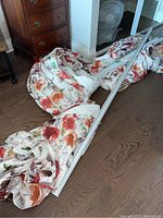 Two pairs of floral lined drapes on the floor next to a dresser, one with longer length than the other, with two white metal curtain rods.