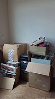 Multiple packed cardboard boxes of assorted CDs and DVDs stacked on a hardwood floor against a wall.