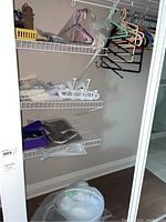 Closet showing mixed wood, plastic, and metal hangers hanging from rod, white plastic over-the-door hooks on side, and 14 inch white table fan on floor.