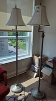 Full view of two metal floor lamps with white fabric shades placed near a window. Lamps are tall, with decorative bases and adjustable swing arms under the shades.