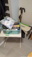 Medical assistance items on white folding table including a plastic tote filled with supplies, N95 masks in zipper bags, and a beige medical stool nearby with multiple walking canes leaning on wall.