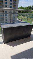 Full view of the Keter outdoor storage box on balcony, showing black resin faux wood texture and horizontal lid.