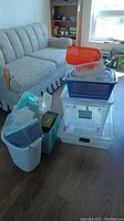 Photo showing a group of various plastic storage containers and baskets stacked and side by side in a room with grey couch and window in background.