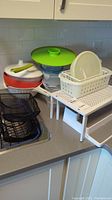 Photo showing shelves, baskets, plastic food containers, salad spinner, mixing bowl with green lid, and colander arranged on kitchen countertop.