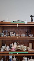 Wooden shelving unit displaying a variety of items including teacups, saucers, plates, cream and sugar bowls, framed carved cork, crystal candle holders, and three desk clocks.