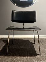 Front view of mid century modern chair showing black patent leather seat and back with chrome legs on carpeted floor against gray wall.