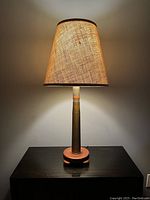 Lamp fully assembled and turned on, showing the light effect of the fabric shade and the artillery shell base on teak wood.
