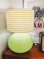 Photo showing the entire lamp with green glass base and white-yellow striped fabric lampshade on a wooden surface next to posters and a radio.