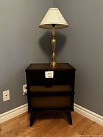 Full view of vintage nightstand with brass table lamp on top against a gray wall.