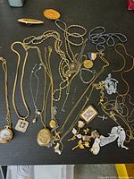 Photo of entire collection showing multiple gold and silver-tone necklaces and pendants along with brooches arranged on a black surface.