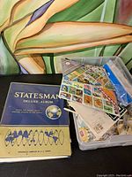 Photo showing the closed Statesman Deluxe Album next to plastic boxes containing loose postage stamps, atop a black surface with a colorful abstract background.