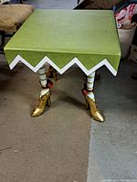 Front and side view of "Elf Legged" side table showing green textured square top with white zigzag trim and decorative elf legs with striped stockings, bows, and gold shoes.