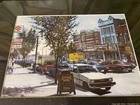 Full view of watercolor painting featuring a street with vintage cars, trees, and storefronts, framed under glass.