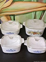 Four square CorningWare saucepans with lids, three with blue cornflower design and one with fruit and vegetable pattern, displayed against a floral backdrop.