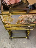 Rack of four folding TV dinner tray tables stacked together showing scenic print tops and gold-tone metal frames with brown wood handles.