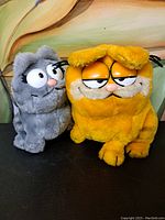 Photo showing Garfield and Nermal plush toys sitting side by side, front view.