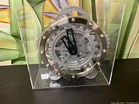 Front view of vintage table clock housed in a clear rectangular enclosure, showing gear-shaped clock face, black hour/minute hands, and surrounding bolts.