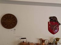 Full photo showing a wooden wall plaque to the left and the Canadian Pacific clock to the right of the plaque on the wall above several metallic pitchers.