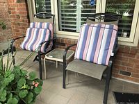 Two metal outdoor chairs with striped cushions and one metal and glass side table between them on a patio. Chairs show wear and scratches.