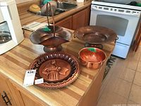 Set of four copper items on kitchen counter: two hammered copper bowls, one copper basket with handle, and one decorative embossed copper plate.
