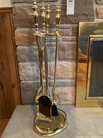 Full view of the brass-finish four-piece fireplace tool set on its circular stand against a stone fireplace background.