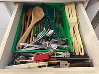 Photo showing a green tray inside a drawer containing various kitchen utensils, spoons, forks, wooden spoons, and spatulas.