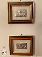 Two smaller framed metal artworks each with wooden and gold-tone frames, light beige matting, featuring rustic landscape scenes with houses and trees in silver metal relief.