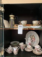 Full view inside cabinet with French press coffee maker, demitasse cups and saucers, tea service pieces including teapot, cream and sugar bowl, decorative plates and black shelving units.