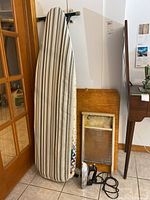 Three vintage laundry items are shown upright against a wall: a folding ironing board with striped cover, a wooden washboard with a discoloration mark on its metal scrubbing surface, and a vintage electric iron with attached cord lying on the floor in front.