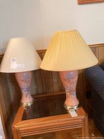 Pair of glass table lamps with different cream-colored shades on wooden table, pink floral bases with silver metal ring bottom.