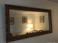 Full view of the rectangular mirror on a wall reflecting a lamp and framed pictures, showing the detailed ornate gilt frame.