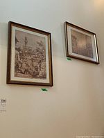 Photo of two framed prints hanging on wall: left is signed, town with tower, right is unsigned street scene.