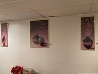 View of three framed prints hanging on a white wall showing minimalist vases with plants in purples and pinks.