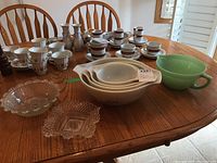 Full view on table showing four white Pyrex mixing bowls nested, green Fire-King Jadeite measuring bowl, two clear pressed glass bowls, and additional tableware in background.
