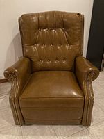 Front view of the brown leather recliner chair showing tufted button backrest, cushioned seat, rolled armrests with metal nailhead trim.