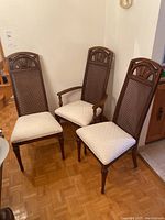 Three cane-backed dining chairs, two armless and one captain chair with arms, showing front and side views, cream cushioned seats, wood frames with carved detail.