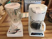 Mr Coffee drip coffee maker and Proctor-Silex blender placed side by side on kitchen countertop. Both appliances are white with glass components and show normal signs of use.