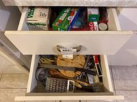 Photo of one drawer open showing various kitchen utensils including a metal grater, ladle, peeler, wooden spoon, and tongs.