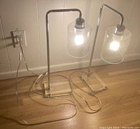 Pair of modern table lamps with chrome arms, clear acrylic rectangular bases, and textured clear glass cylindrical shades illuminated with bulbs.