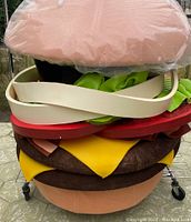Full stacked view showing hamburger necklace layers: bun, lettuce, tomato, bacon, cheese, burger patties, bottom bun, on rolling cart.