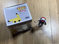 Photo of Pikachu Funko Pop box next to Ash figurine on wood floor