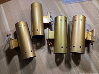 Four gold cylindrical Hollywood Regency style wall sconces placed on a wooden floor. Each has electrical wiring and mounting brackets visible. Surface shows some wear and scratches.