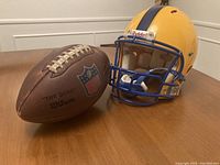Photo of Wilson NFL 'The Duke' football next to Riddell football helmet, displaying ball's NFL shield logo and leather texture, and helmet's profile showing yellow shell and blue facemask.